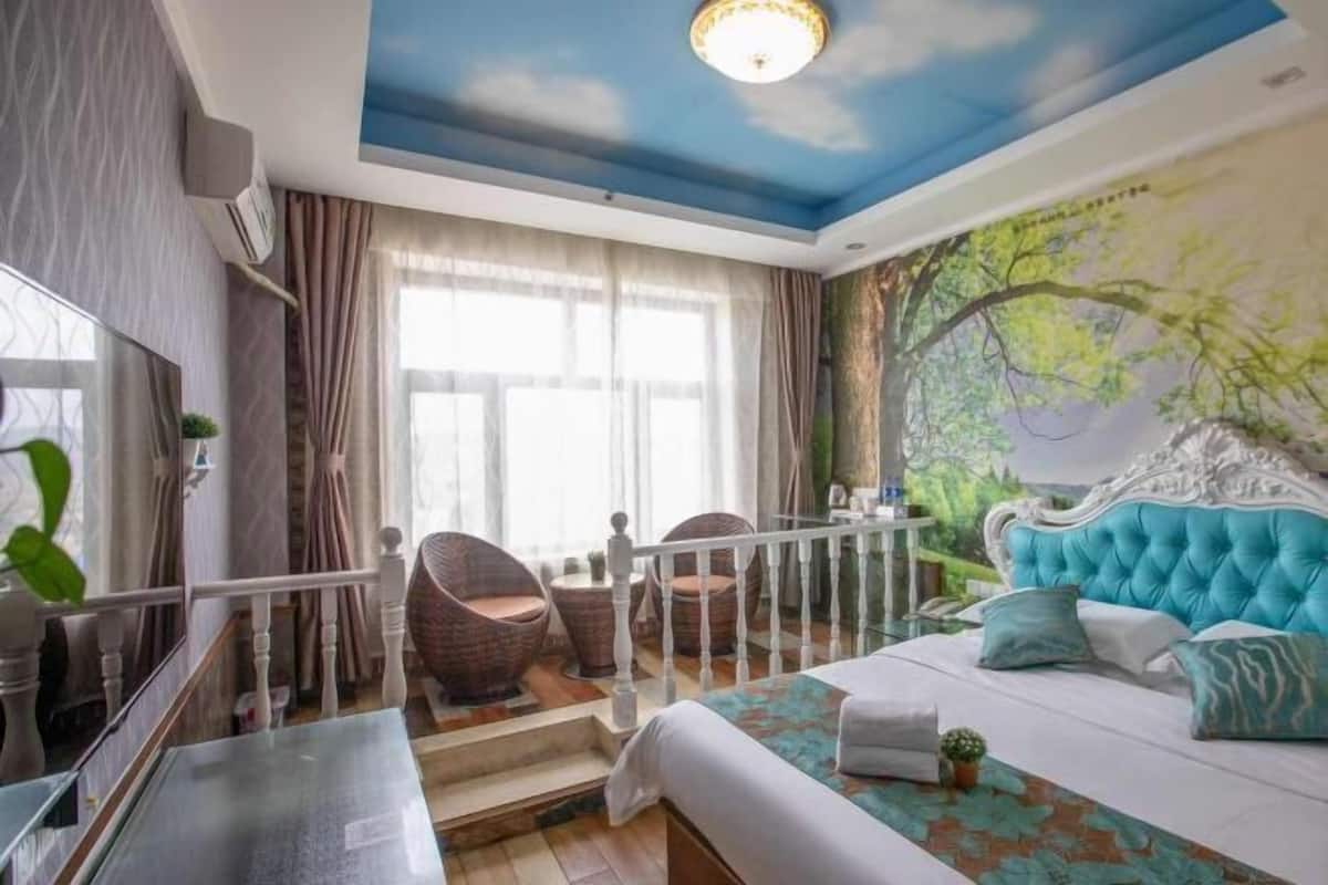 Une chambre à coucher avec un lit, une peinture murale au plafond, un escalier, des chaises en osier et une vue sur la verdure à l’extérieur.