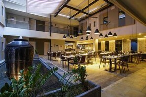 A spacious indoor dining area with multiple tables and chairs, a large planter with plants, and hanging pendant lights. - Sevn Legian (Bali)