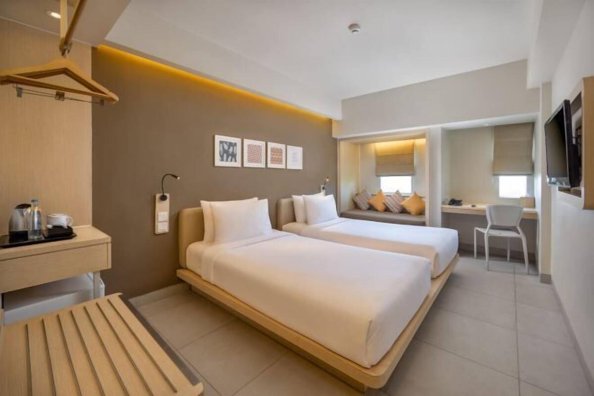A hotel room with two beds, a desk, a chair, a TV, and a small kitchenette.