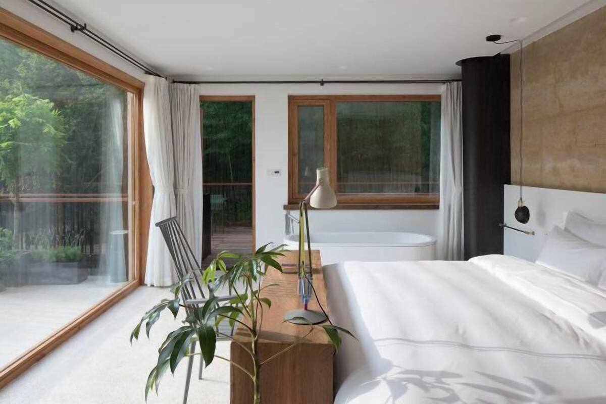 A modern bedroom with a large bed, a wooden nightstand, a potted plant, and a view of a balcony.