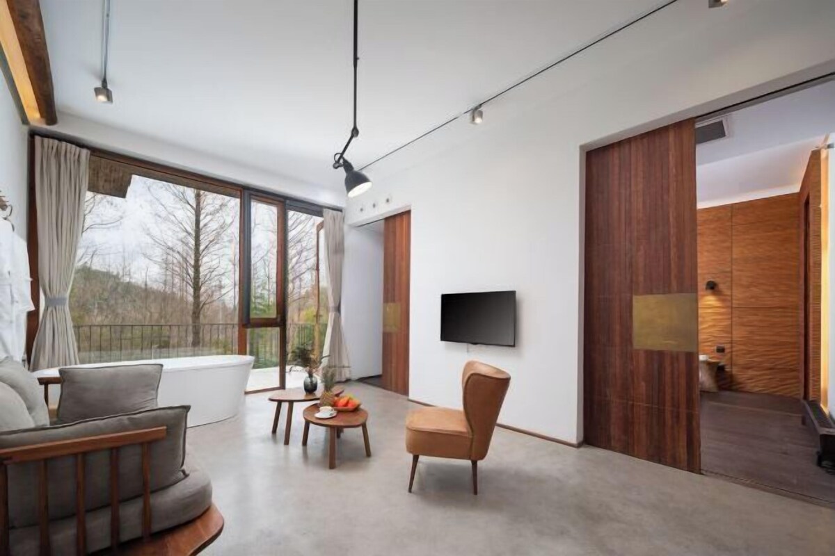 A modern living room with a sofa, armchair, and coffee table, a TV mounted on the wall, and a large window with curtains.