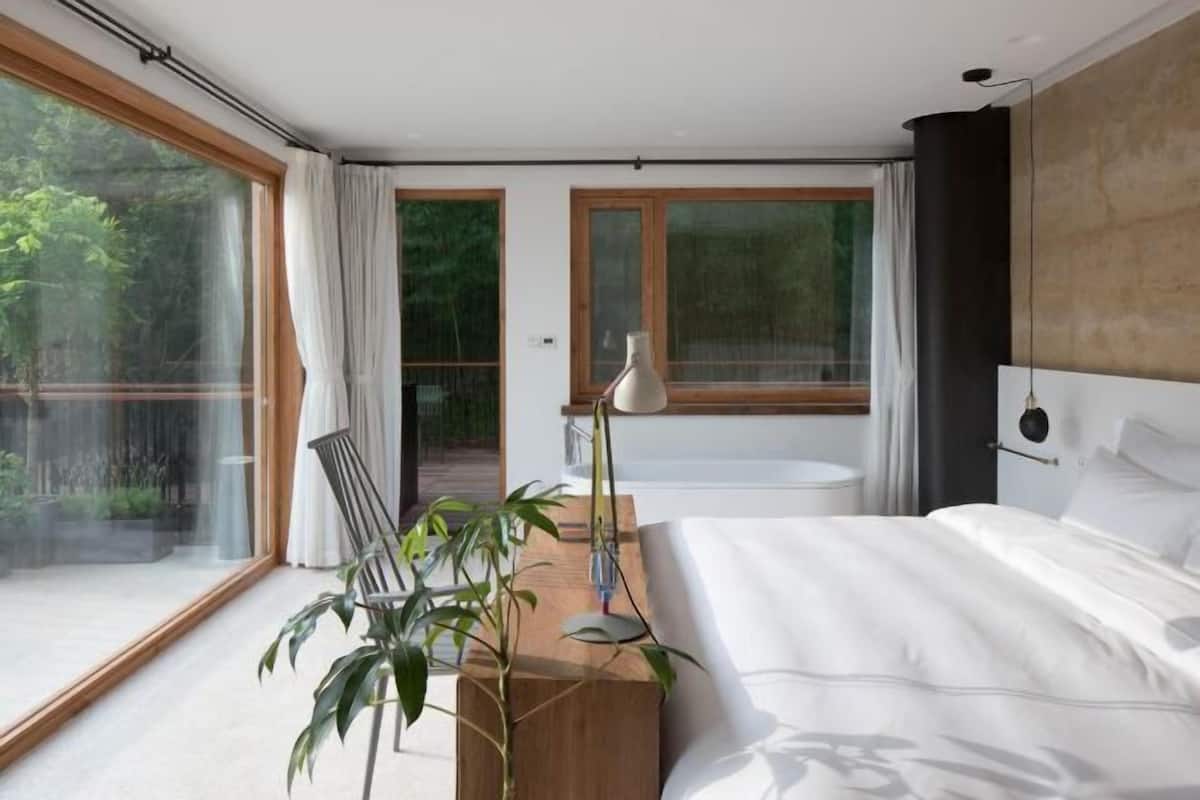 A modern bedroom with a large bed, a wooden side table, a floor lamp, and a view of the outdoors through large windows.