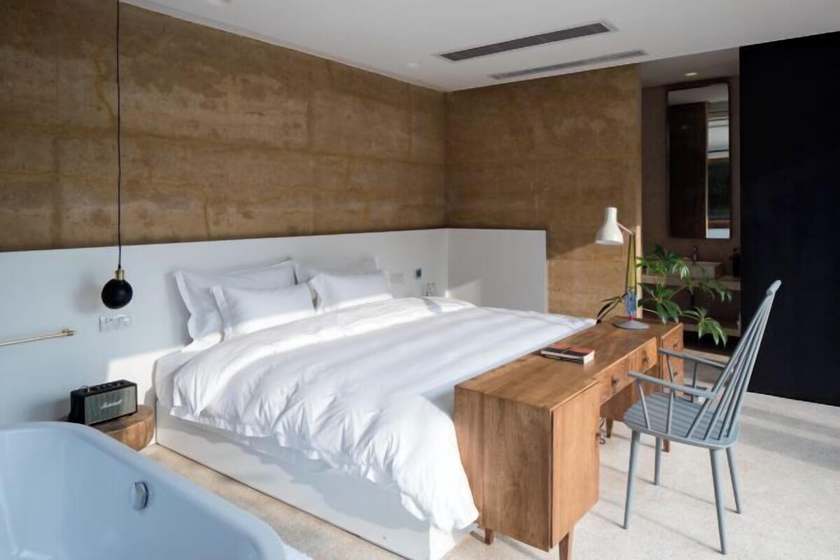 A modern bedroom with a large bed, a wooden desk, a chair, a bedside table, a lamp, and a bathtub.