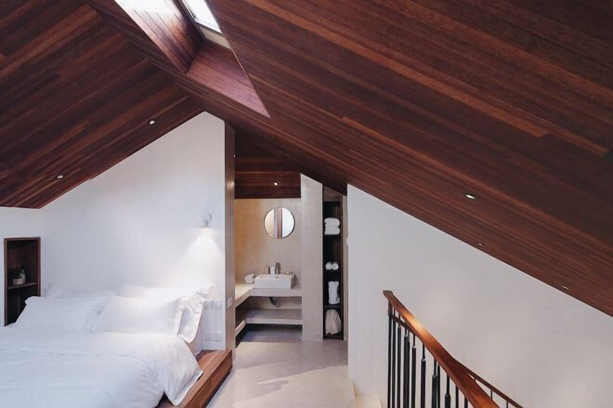 A bedroom with a sloped ceiling, a bed, a bedside table, a mirror, and a bathroom with a sink and towel rack.