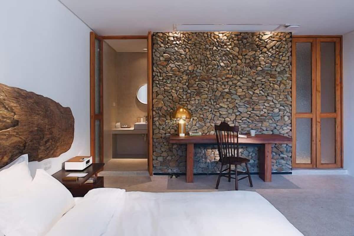 A bedroom with a large bed, a wooden nightstand, a desk with a chair, and a stone wall.