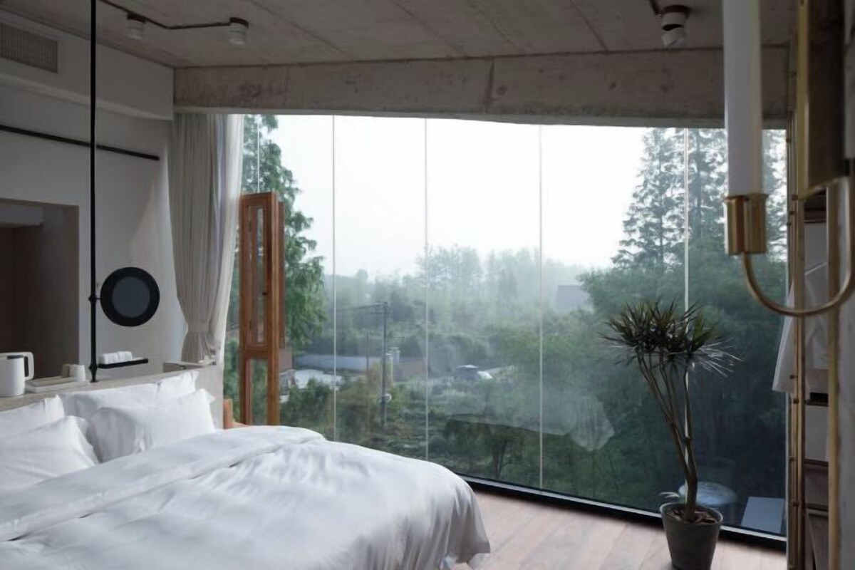 A bedroom with a large window, a bed, a bedside table, and a potted plant.