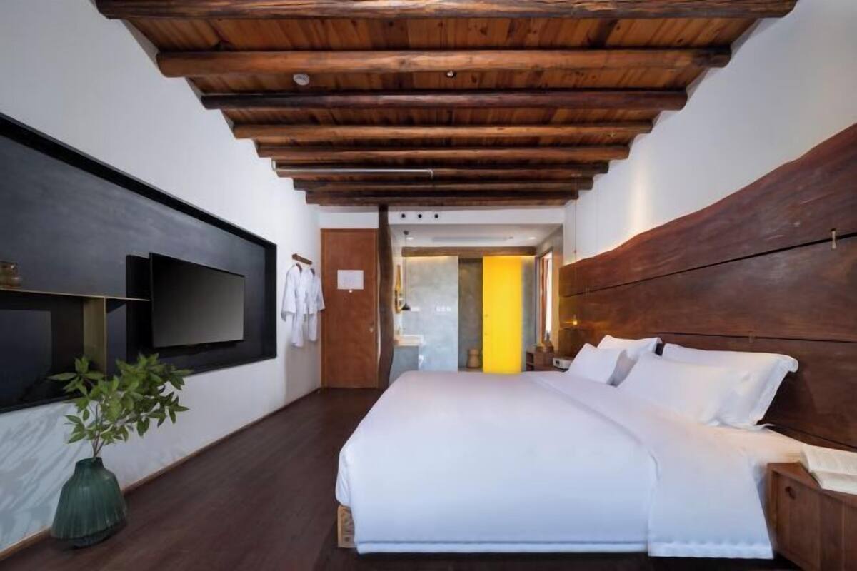A bedroom with a wooden ceiling, a bed with white linens, a bedside table, a wall-mounted TV, and a potted plant.