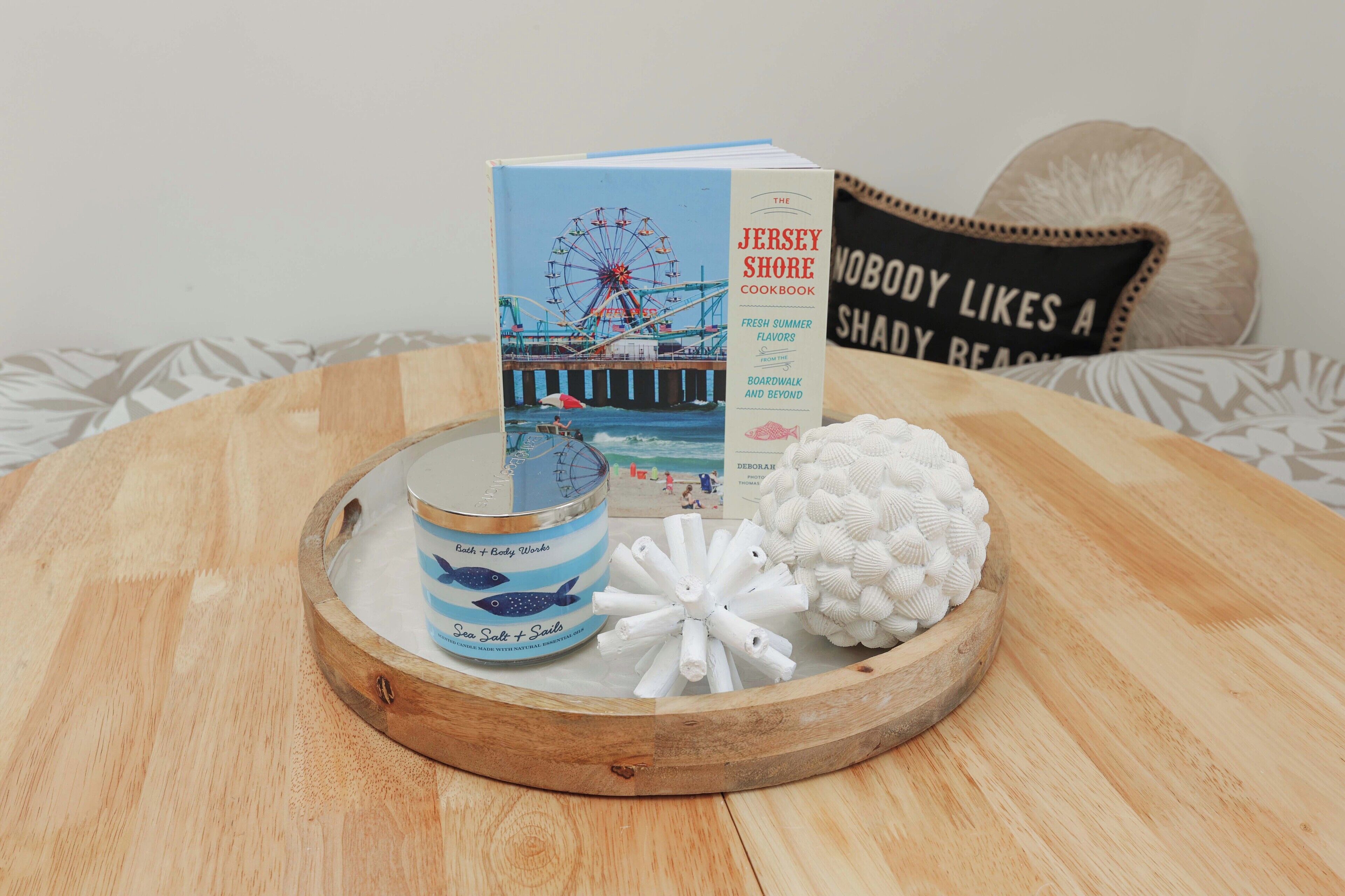 A wooden tray with a book titled "The Jersey Shore Cookbook," a candle, and decorative items on a patterned fabric surface.