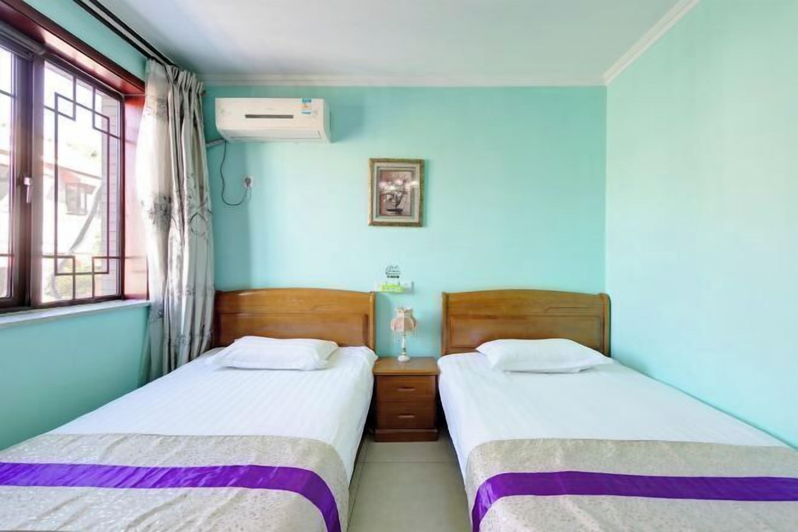 A room with two beds, a window with curtains, an air conditioning unit, and a small bedside table with a lamp.