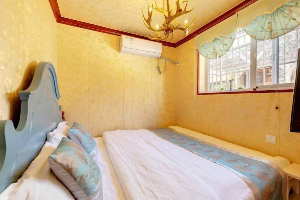 A small, single-bed room with a wooden headboard, a window with a curtain, and a chandelier.