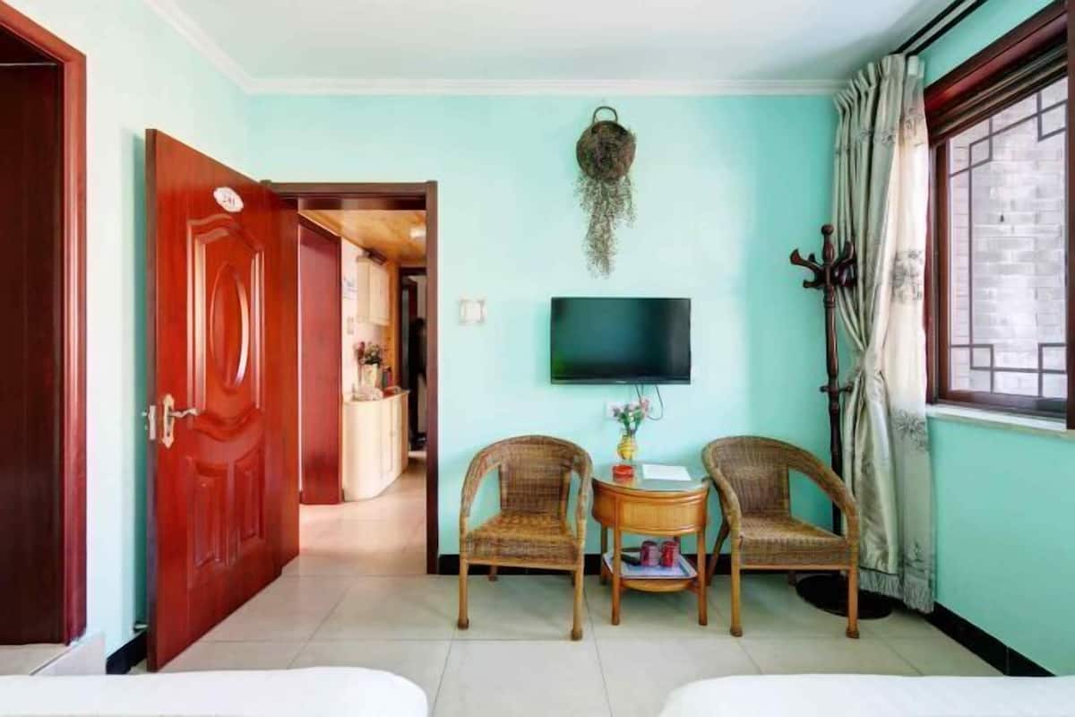 A room with two wicker chairs, a small table with a vase, a flat-screen TV mounted on the wall, and a window with curtains.