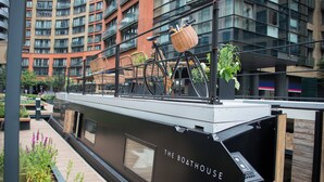 Ein modernes Boot mit der Aufschrift "THE BOATHOUSE", ausgestattet mit einem Fahrradträger und einem Korb, liegt in einem städtischen Uferbereich vor dem Hintergrund hoher Gebäude.