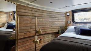 Ein gemütliches Interieur mit Holzwandverkleidung, einem Bett mit dunklem Kopfteil sowie einem Nachttisch mit Lampe und kleiner Pflanze.