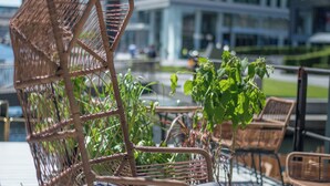 Ein Korbstuhl mit Kissen, ein Blumentopf und ein Glastisch auf einer Terrasses mit Blick auf moderne Gebäude.