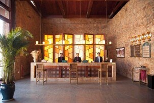 Zona de recepción con mostrador de madera, una planta en maceta grande y una pared de piedra con un vitral.