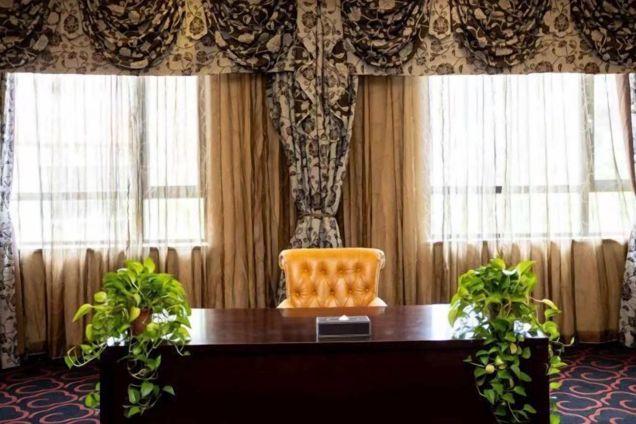 A reception desk with a tufted chair, two potted plants, and patterned curtains.