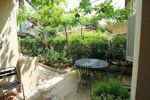 A courtyard with a round table and chairs, surrounded by greenery and a building in the background.