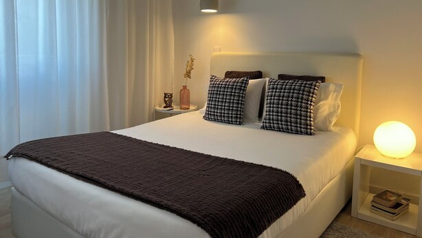 A neatly made bed with a brown blanket, two patterned pillows, and a bedside lamp. - Burgães II Apartment (Porto)