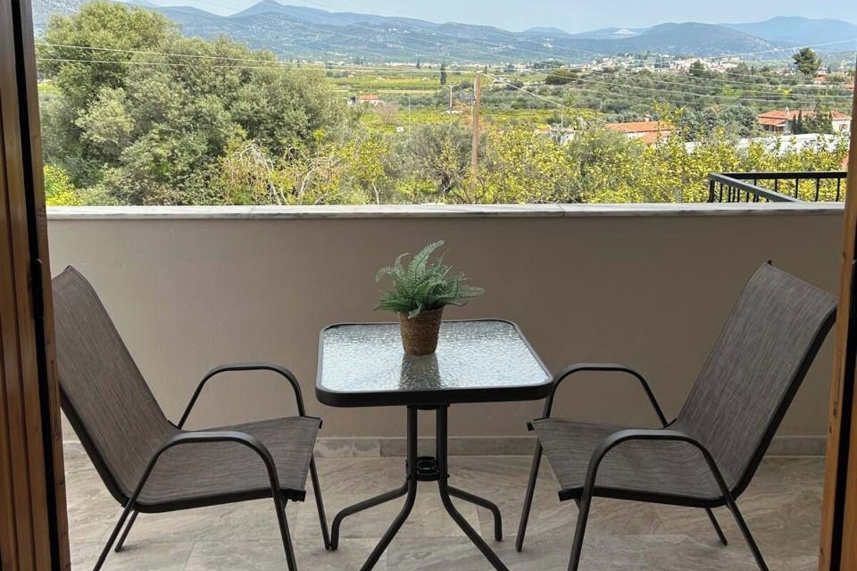 A balcony with a table and two chairs, a potted plant, and a view of a landscape with trees and distant hills.