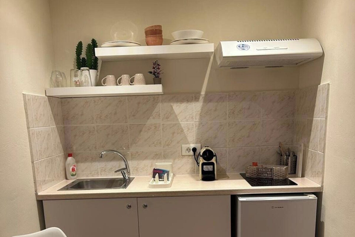 A compact kitchen with a sink, a small refrigerator, a chair, and a shelf with dishes and a kettle.