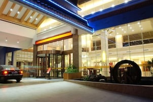 Entrada de un hotel con fachada de vidrio, puerta giratoria y una gran escultura circular al frente.