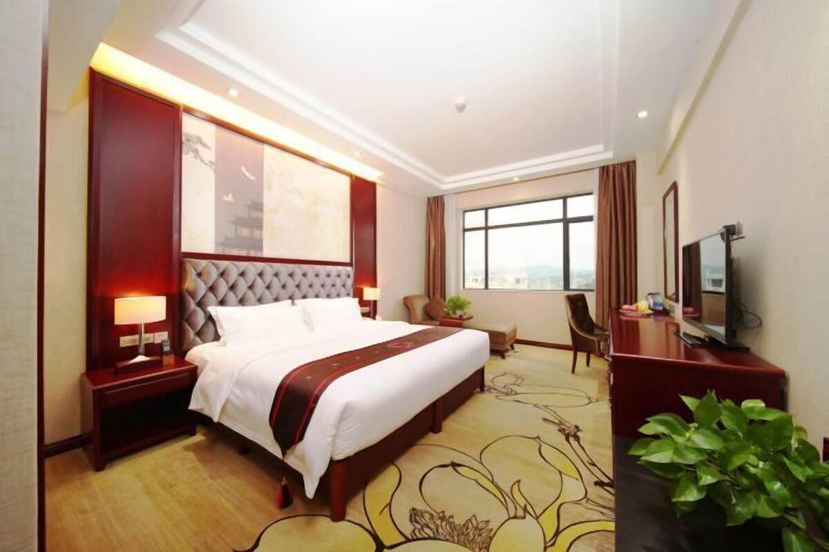 Habitación de hotel con una cama grande, un escritorio con televisor, una ventana con vistas a la ciudad y un panel decorativo en la pared.