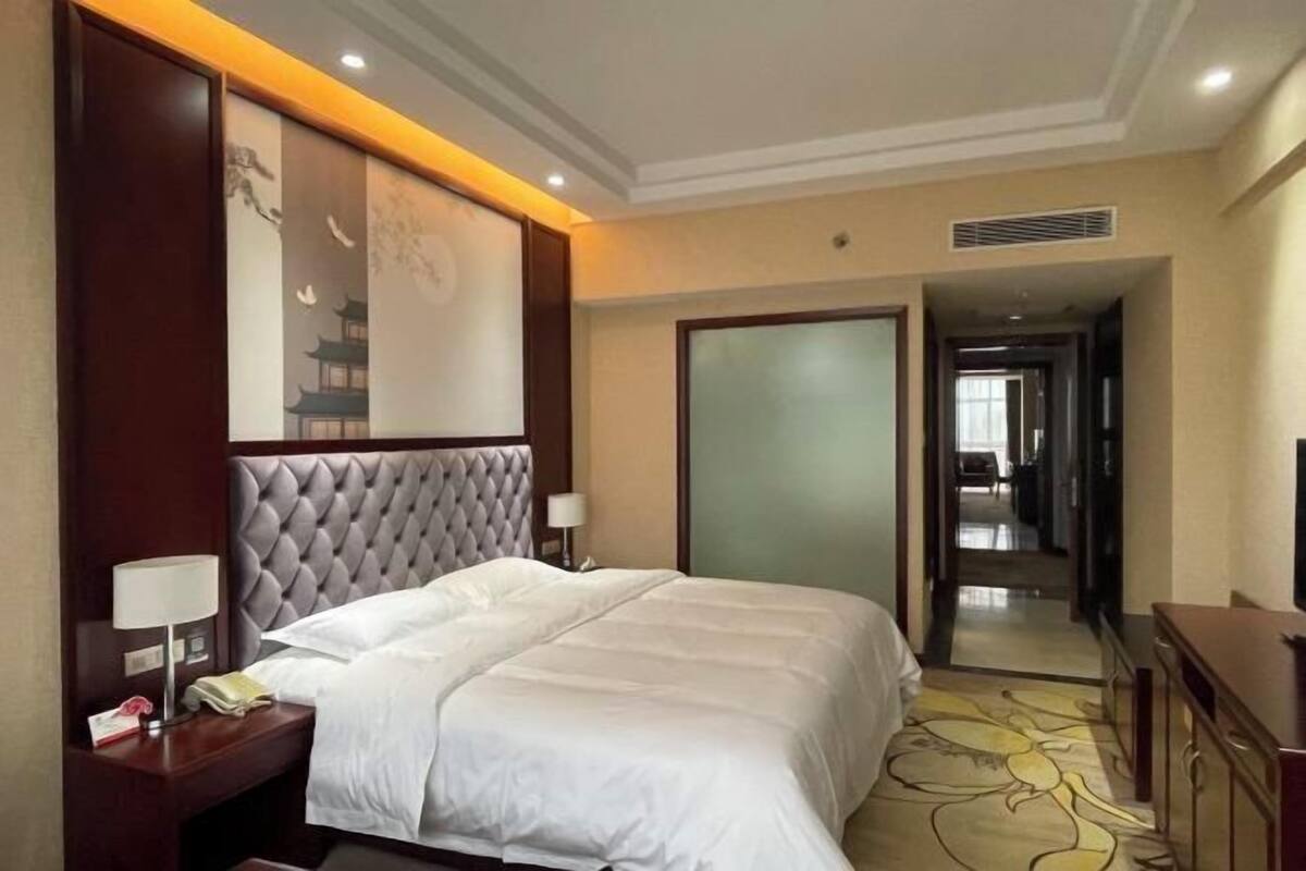 Habitación de hotel con una cama grande, una mesita de noche con lámpara, una obra de arte en la pared y una puerta que da a otra habitación.