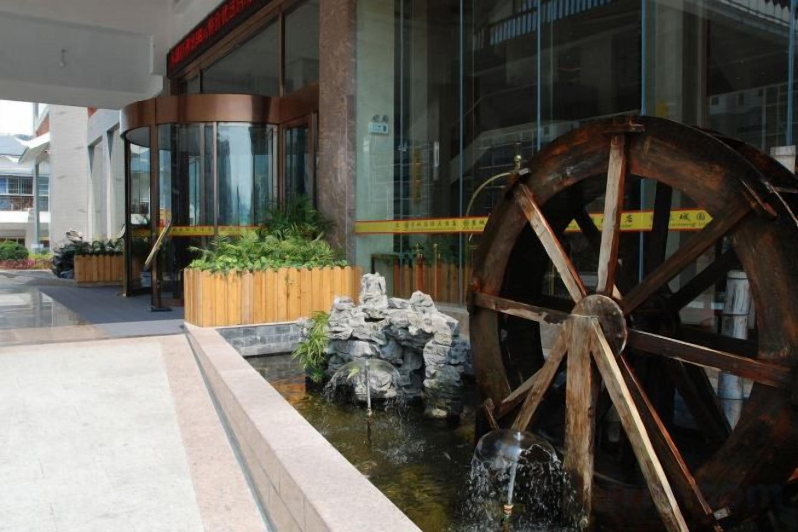 A building with a large wooden water wheel, a stone fountain, and a wooden planter with greenery.