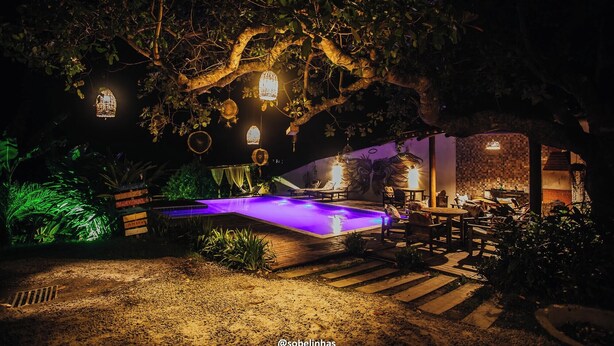 A nighttime outdoor pool area with a wooden deck, lounge chairs, and hanging lanterns. - Villa Nobre Trancoso (Porto Seguro)