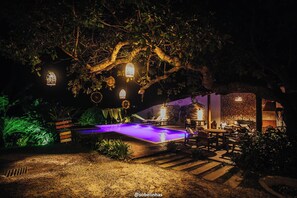 A nighttime outdoor pool area with a wooden deck, lounge chairs, and hanging lanterns. - Villa Nobre Trancoso (Porto Seguro)
