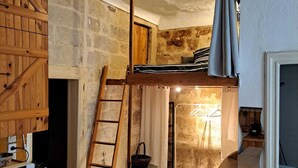 A rustic room with a loft bed, wooden furniture, a stone wall, and a small table with a cup.