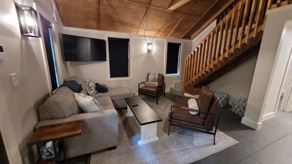 Una sala de estar con sofá, mesa de centro, sillón, una escalera y un televisor montado en la pared.