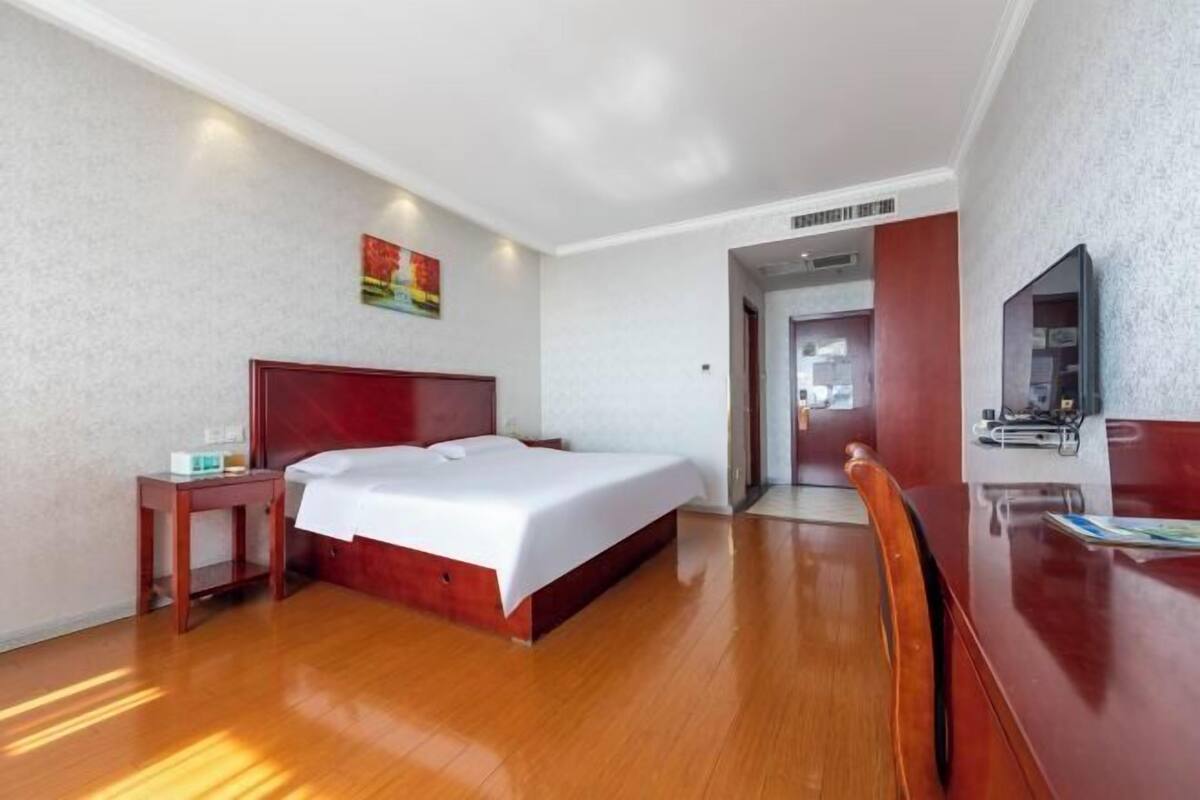 A hotel room with a wooden bed, a bedside table, a chair, a TV, and a door leading to another room.