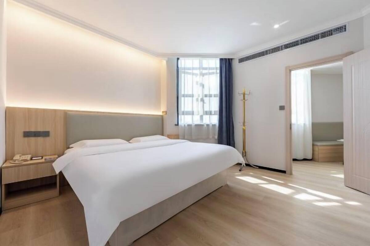 A modern hotel room with a large bed, bedside table, lamp, and a view of the outdoors.