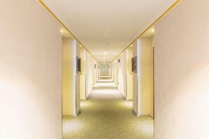Un long couloir étroit, typique des hôtels, avec un tapis à motifs, des murs de couleur claire et des portes disposées à intervalles réguliers.