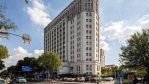 Un edificio alto y rectangular con varios pisos y techo plano, rodeado de árboles y automóviles estacionados.