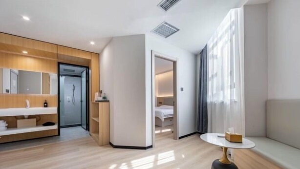 Habitación de hotel moderna con suelo de madera, una mesa redonda blanca, sofá, armario empotrado y zona de ducha.