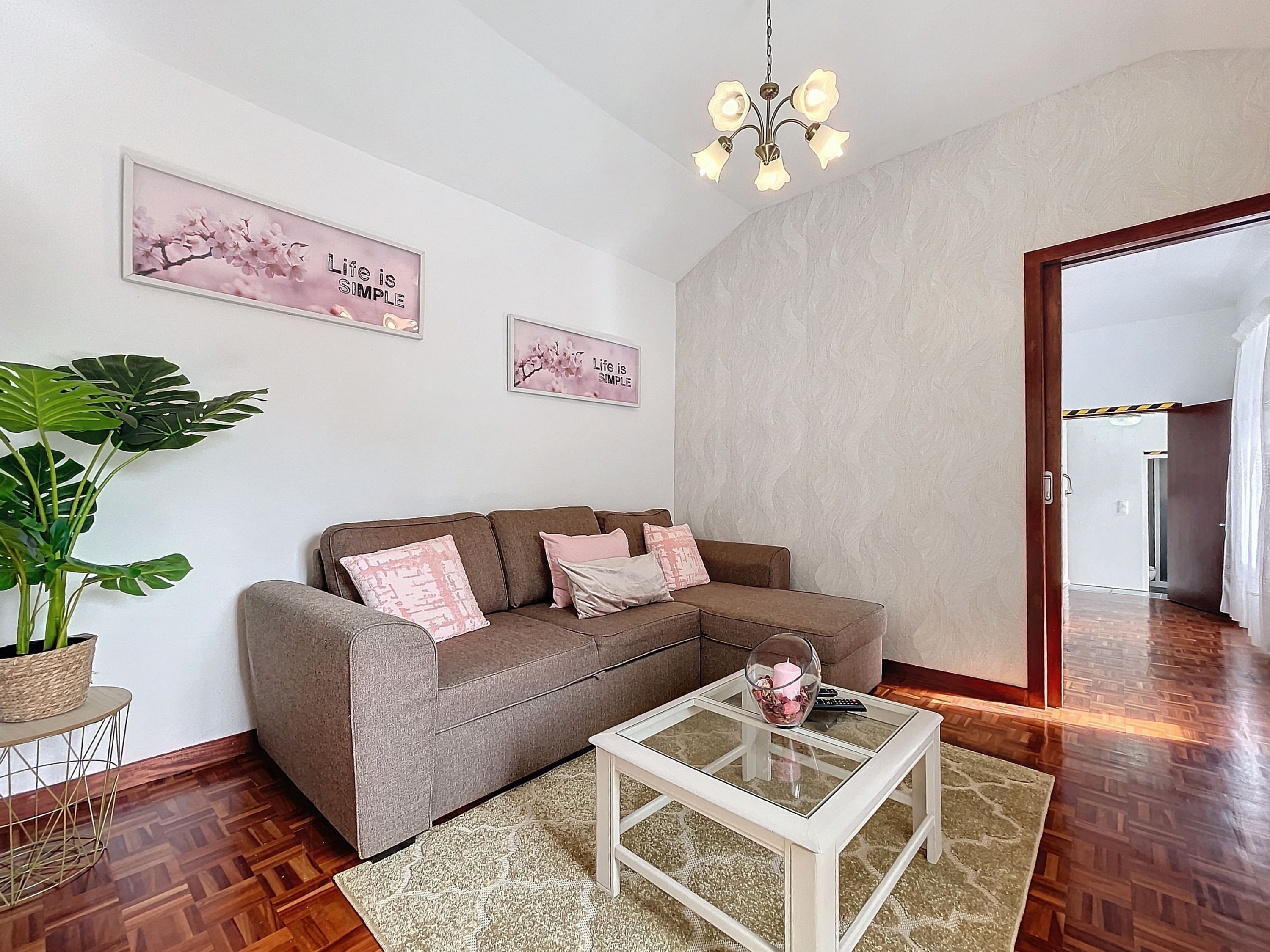 A living room with a brown sofa, a glass coffee table, a potted plant, and two wall art pieces.
