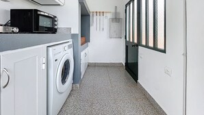 Lavandería con lavadora, secadora y microondas. Hay una ventana con persianas y una puerta con picaporte.
