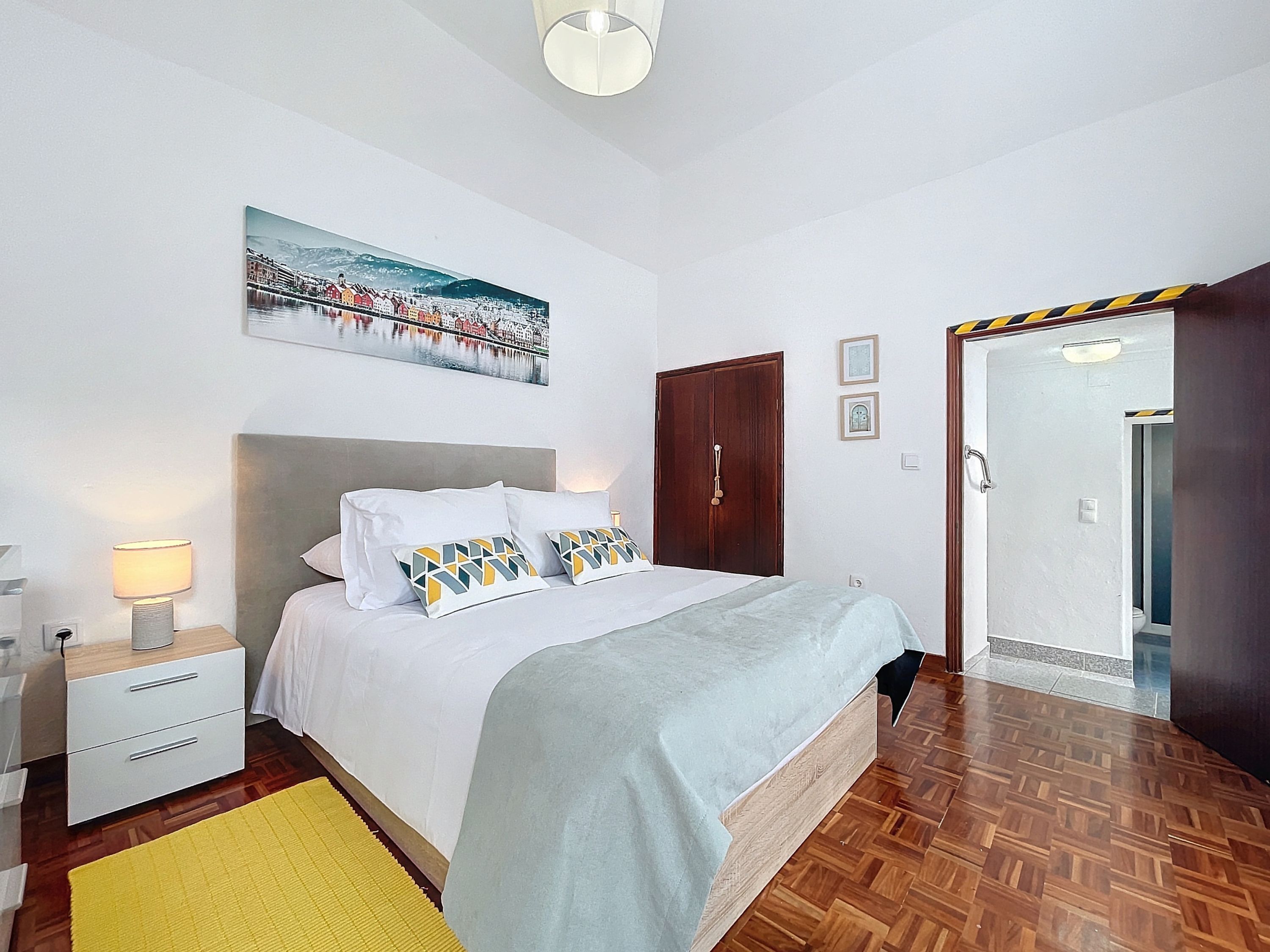 A bedroom with a bed, bedside tables, a lamp, a painting, a wooden door, and a bathroom visible through an open door.