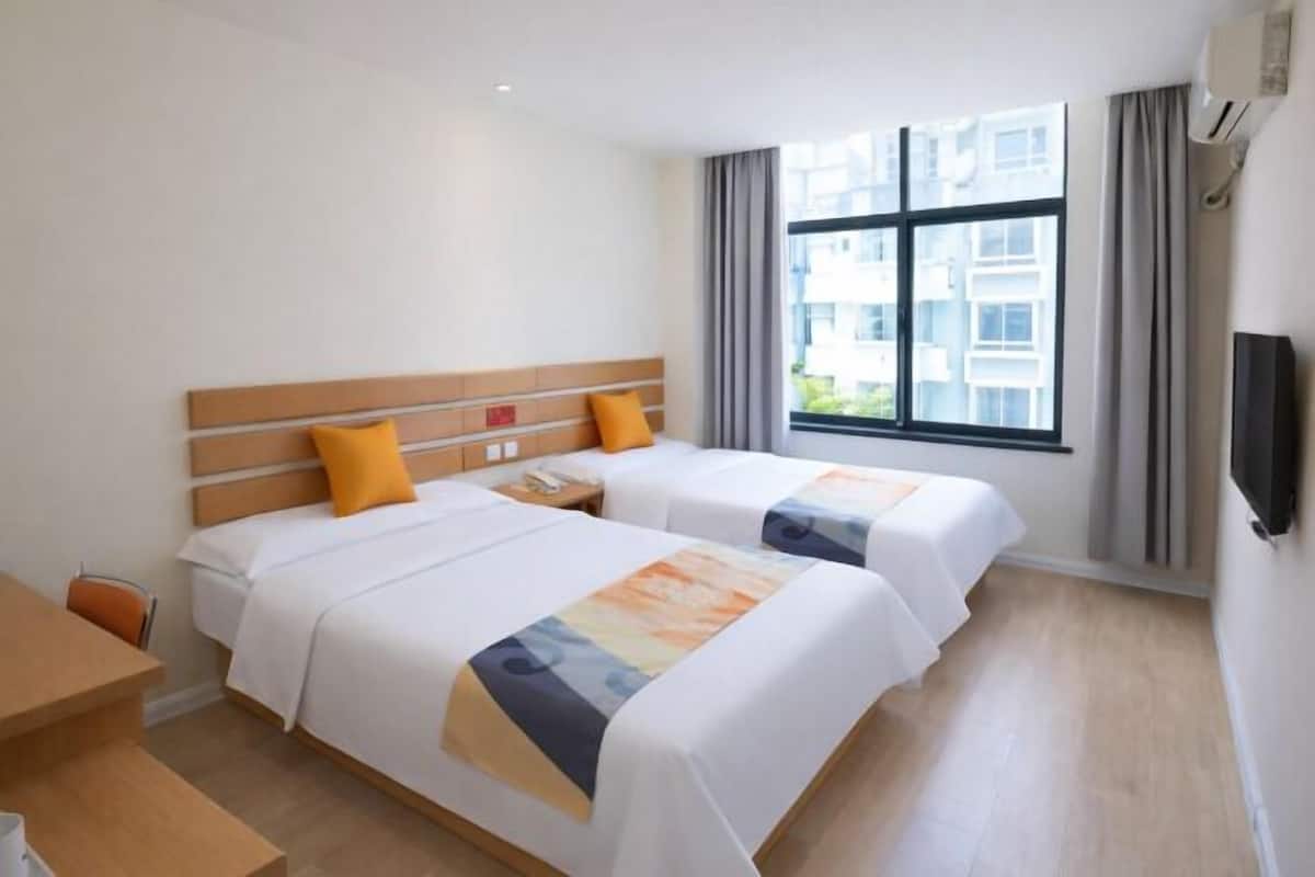 Une chambre d’hôtel avec deux lits, une tête de lit en bois, une fenêtre avec des rideaux et un téléviseur à écran plat fixé au mur.