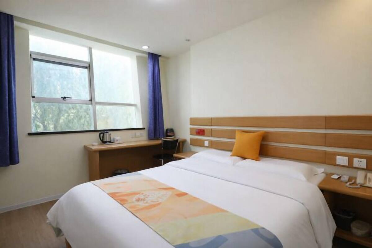 Une chambre d’hôtel comprenant un lit, un bureau, une chaise, une fenêtre avec des rideaux et une table de chevet avec une lampe.