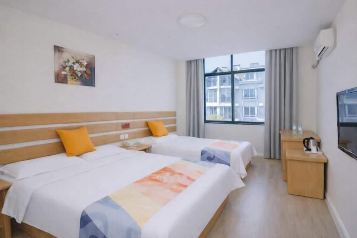Une chambre d’hôtel avec deux lits, une tête de lit en bois, un tableau au mur, une fenêtre avec des rideaux, un bureau avec une bouilloire et une télévision.