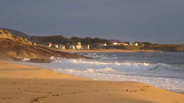 A coastal beach scene with a rocky shoreline, waves, and a row of houses in the background.