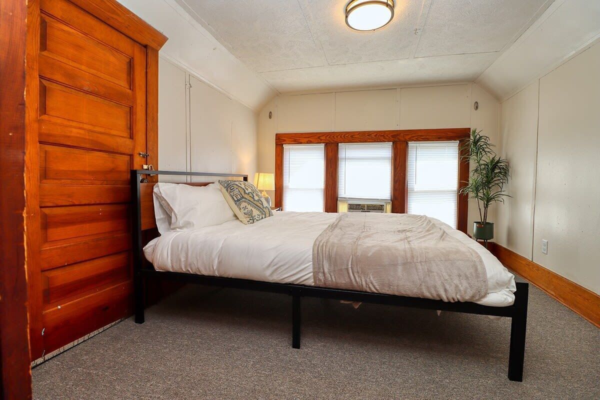 A single bed with a wooden frame, white bedding, and a bedside lamp. There is a wooden door to the left and a window with blinds to the right.