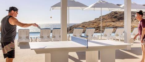 A table tennis table with chairs and umbrellas in a sunny outdoor area with a view of the sea.