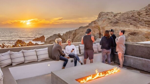 A group of people seated on a modern outdoor lounge area with a fire feature, overlooking a rocky coastline at sunset. - Oceanfront 2BR w/ Two King Beds • Cerritos Beach (Los Cerritos)