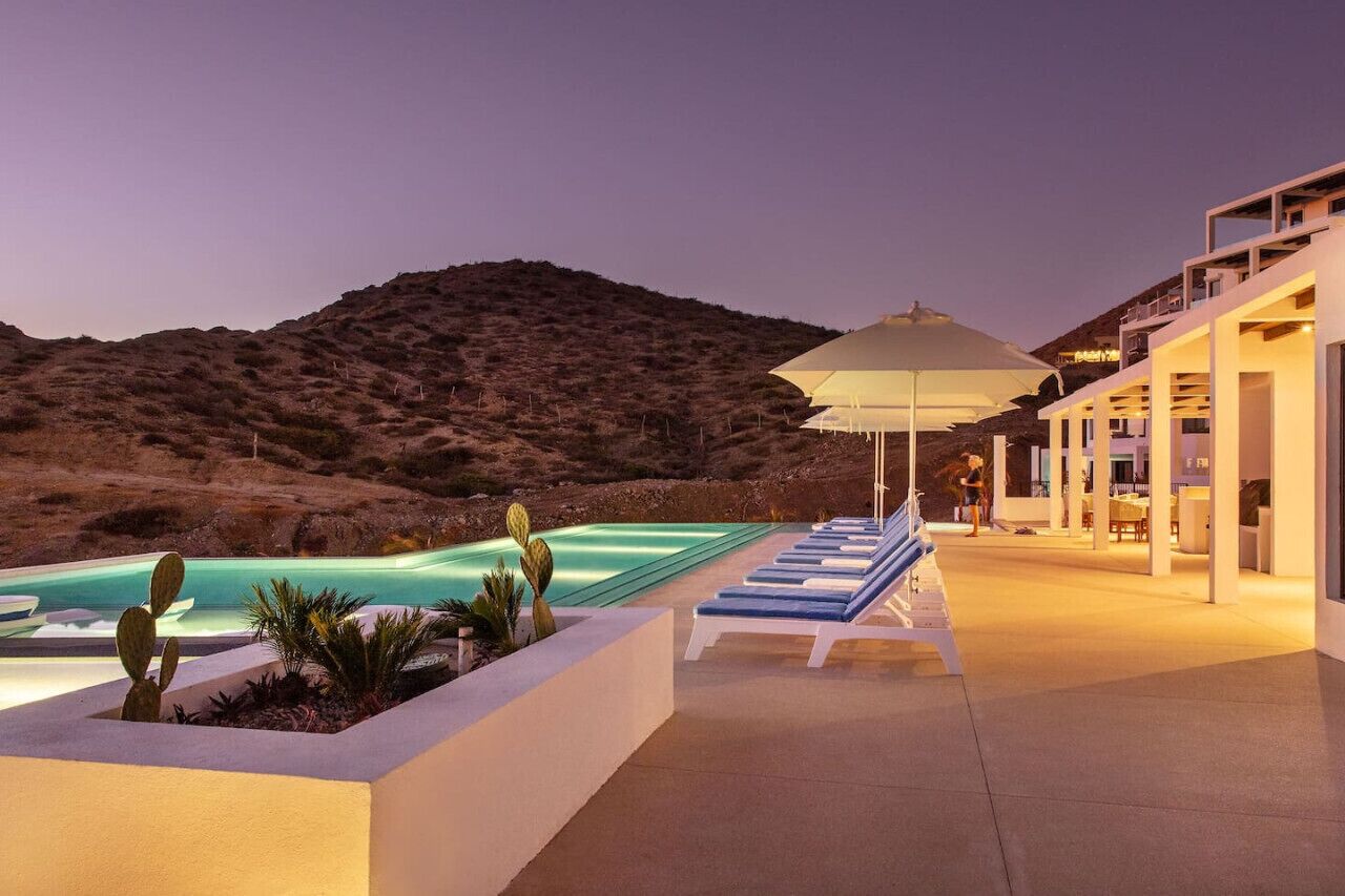Un espace piscine doté de chaises longues et d’un grand parasol, entouré d’un paysage désertique et d’un bâtiment comportant de nombreux balcons.