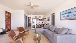 A modern living room with a grey sofa, wooden chairs, a glass coffee table, and a ceiling fan. - Oceanfront 2BR w/ Two King Beds • Cerritos Beach (Los Cerritos)