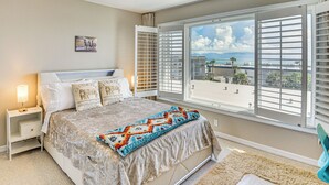 Een slaapkamer met een bed, nachtkastje, raam met jaloezieën, plafondventilator en uitzicht op het strand.
